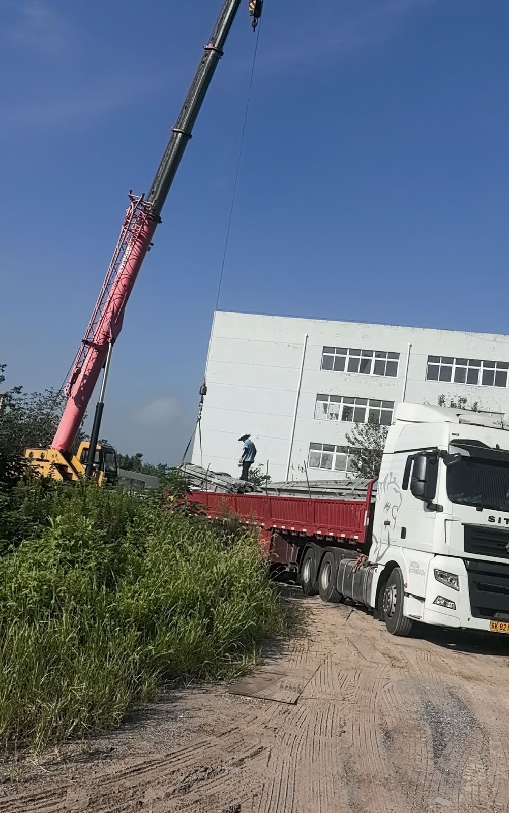 吊车正在往货车上吊装二手钢结构构件，红色半挂车停在货场